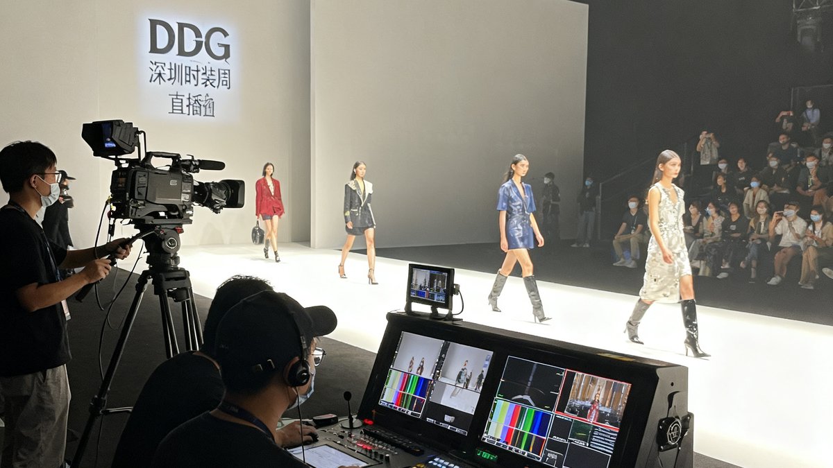 深圳时装周官方直播现场，DG视讯团队操作着多台摄像机与专业导播台，T台上模特正在走秀